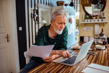 Older man sits at kitchen counter reviewing papers and laptop