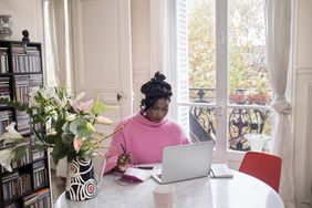 person in bright pink sweater writing something in notebook in a sunny room