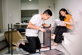 A couple in a living room putting together a desk with one partner reading the instructions and the other doing the assembly 