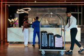A man and woman check in at hotel front desk while a worker wheels a luggage cart through the lobby