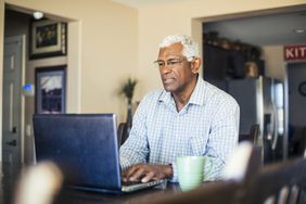 An older man sits on a laptop at his table at home