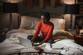 Young woman sitting on bed uses laptop in evening