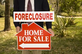 Realtor holds foreclosure for sale sign in the front yard of a home.