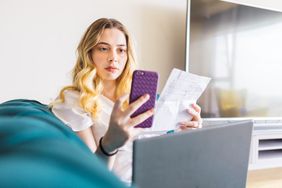 surprised and confused young woman checking her bank balance on her smartphone while holding a student loan bill