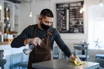 Young man barista with face mask and gloves standing in coffee shop, disinfecting tables.