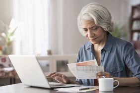 Senior woman examining paperwork at table at home with laptop