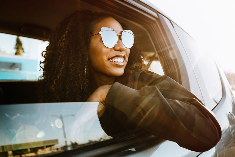 Young woman wearing sunglasses smiling with arm out car window