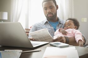 Father holding sleeping baby while doing paperwork at the computer