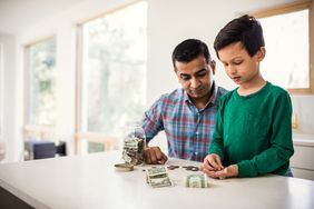 A parent and young child count money on a kitchen table at home