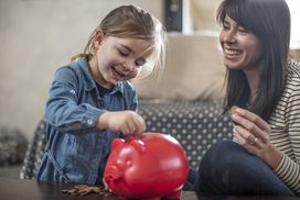 Girl inserting coin from mother into red piggy bank on coffee table