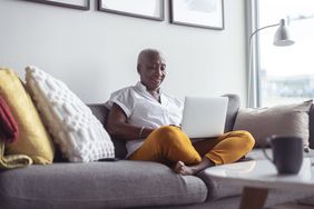 A homeowner sits comfortably on a sofa looking at reverse mortgage requirements on a laptop.