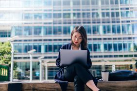 Woman with laptop sits on bench outside office building