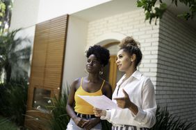 A woman and a real estate agent holding a document have a discussion in front of a home