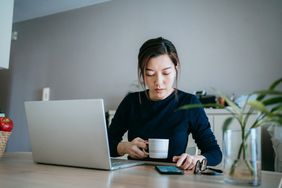 Young woman works on laptop and cellphone while drinking coffee