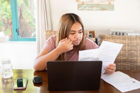 Woman at kitchen table reviews paper documents with laptop open