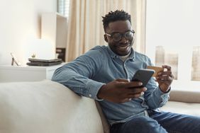 Man sits on couch with a smartphone and credit card in hand, paying for something he bought online