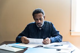 An older man sits at a table, frowning as he looks at a pile of tax paperwork spread out in front of him