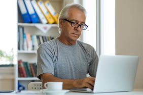 Silver haired bespectacled man working on laptop with white coffee mug on table