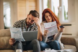 Worried couple seated on a couch looking over their paper bills; laptop on man^as lap