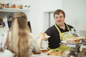 A person with Down syndrome serving a scone to a woman in a cafe where he works