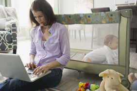 Woman on laptop sits on floor in front of baby in playpen