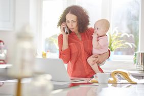 Woman holding baby talks on landline and looks at laptop