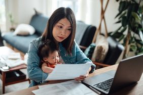 Parent holding a baby and working with papers and a laptop