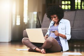 Black woman sits on hardwood floor in front of a couch with a laptop and a credit card in her hand. 