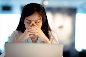 A woman behind a laptop screen rests her eyes with a worried expression.