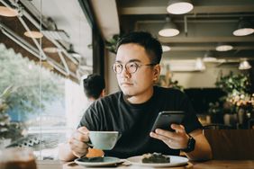 A young man sits at a cafe table and stres out the window to his right. He holds a cup of tea in his right hand and a smartphone in his left.