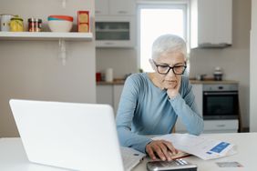 Senior woman working on a calculator at her kitchen table with laptop and documents nearby